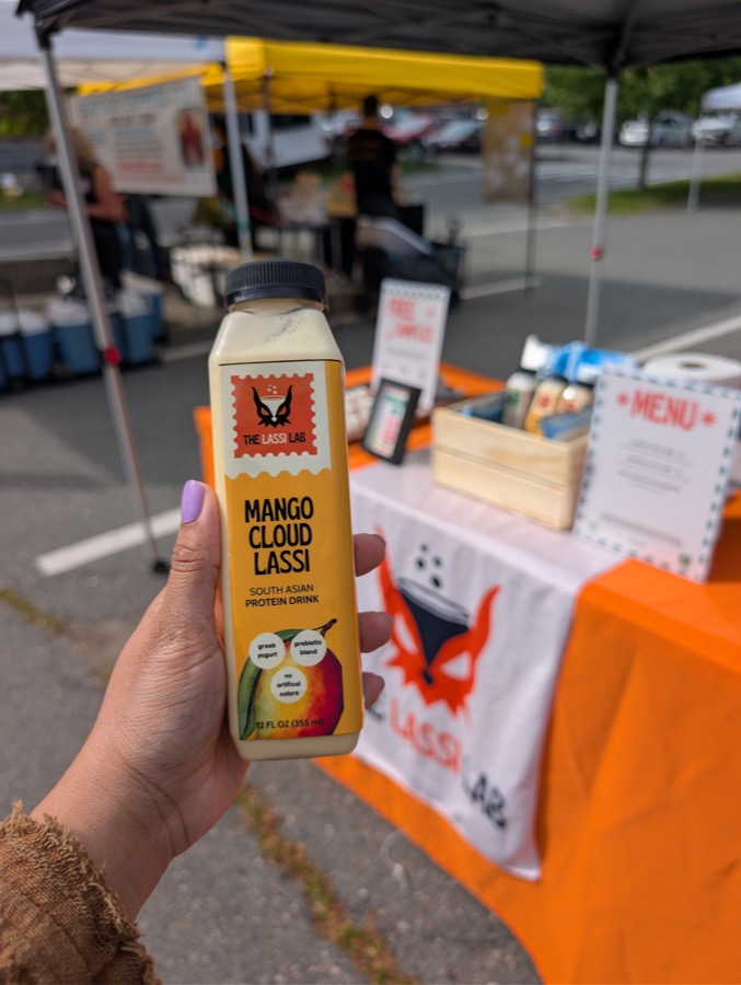 A hand holding a Mango Cloud Lassi bottle in front of the market tent